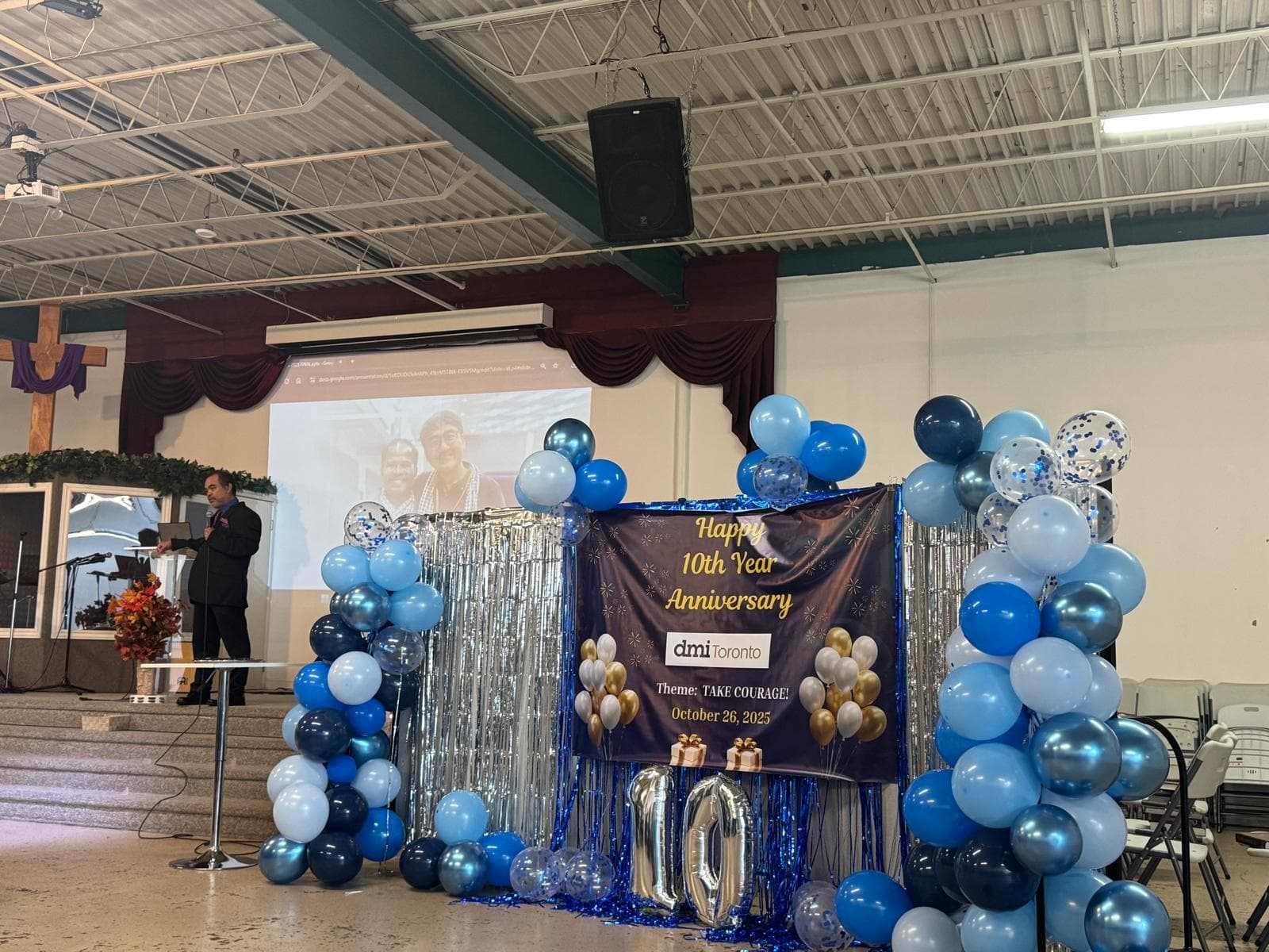 A stage decorated with a Take Courage banner, a giant silver number 10, and blue and silver balloons.